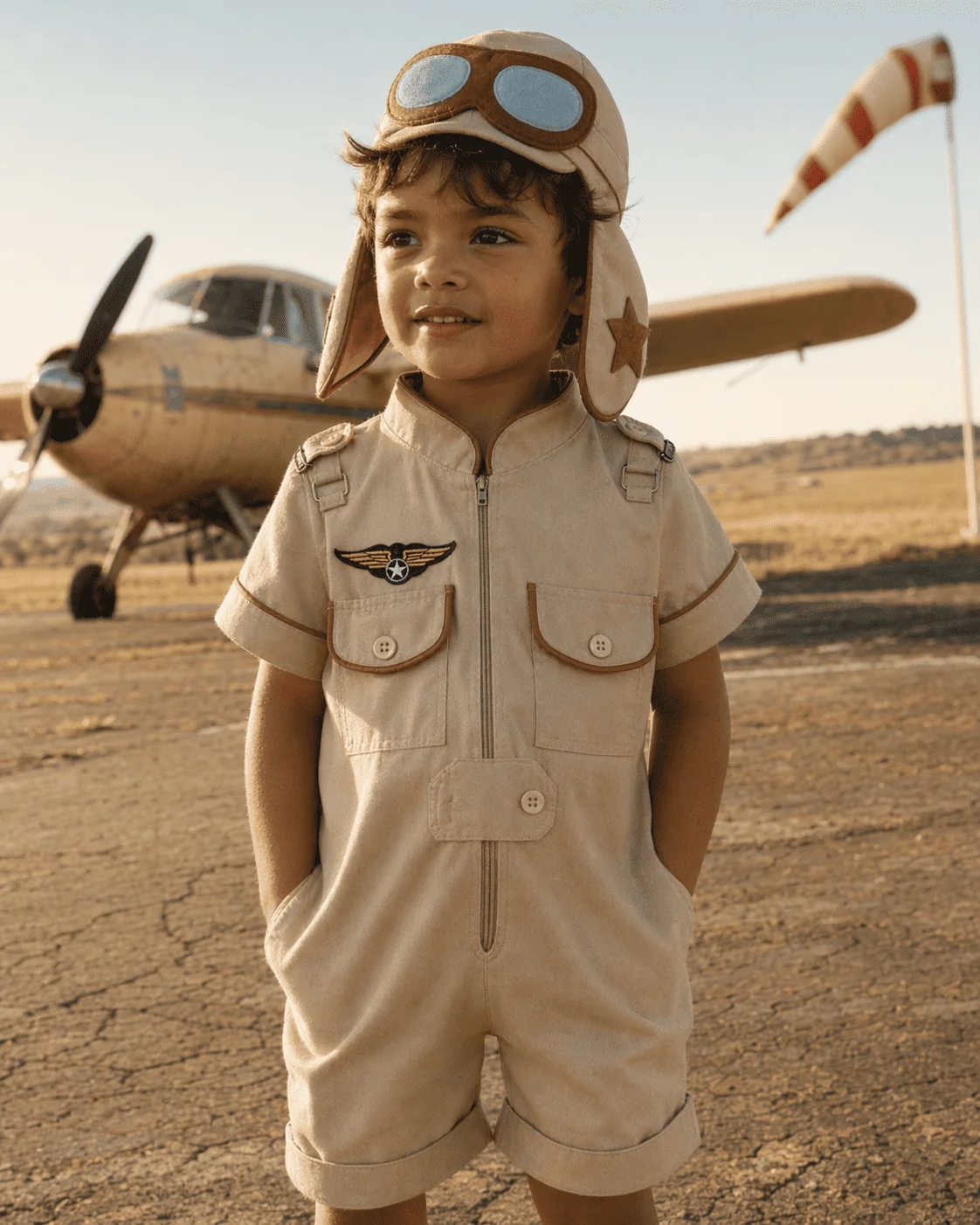 Menino brasileiro sorrindo em macacão Aviador curto sarja bege na pista do aeroclube — touca de aviador com óculos, bordado de asas douradas no peito, viés caramelo nos bolsos, avião vintage e biruta ao fundo