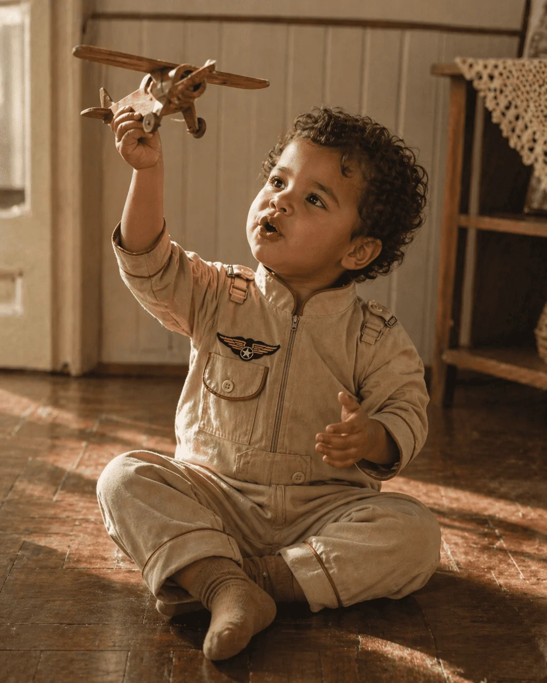 Toddler brasileiro sentado no chão de madeira brincando com aviãozinho de madeira, vestindo Aviador longo sarja bege com bordado de asas no peito, luz natural ambarina