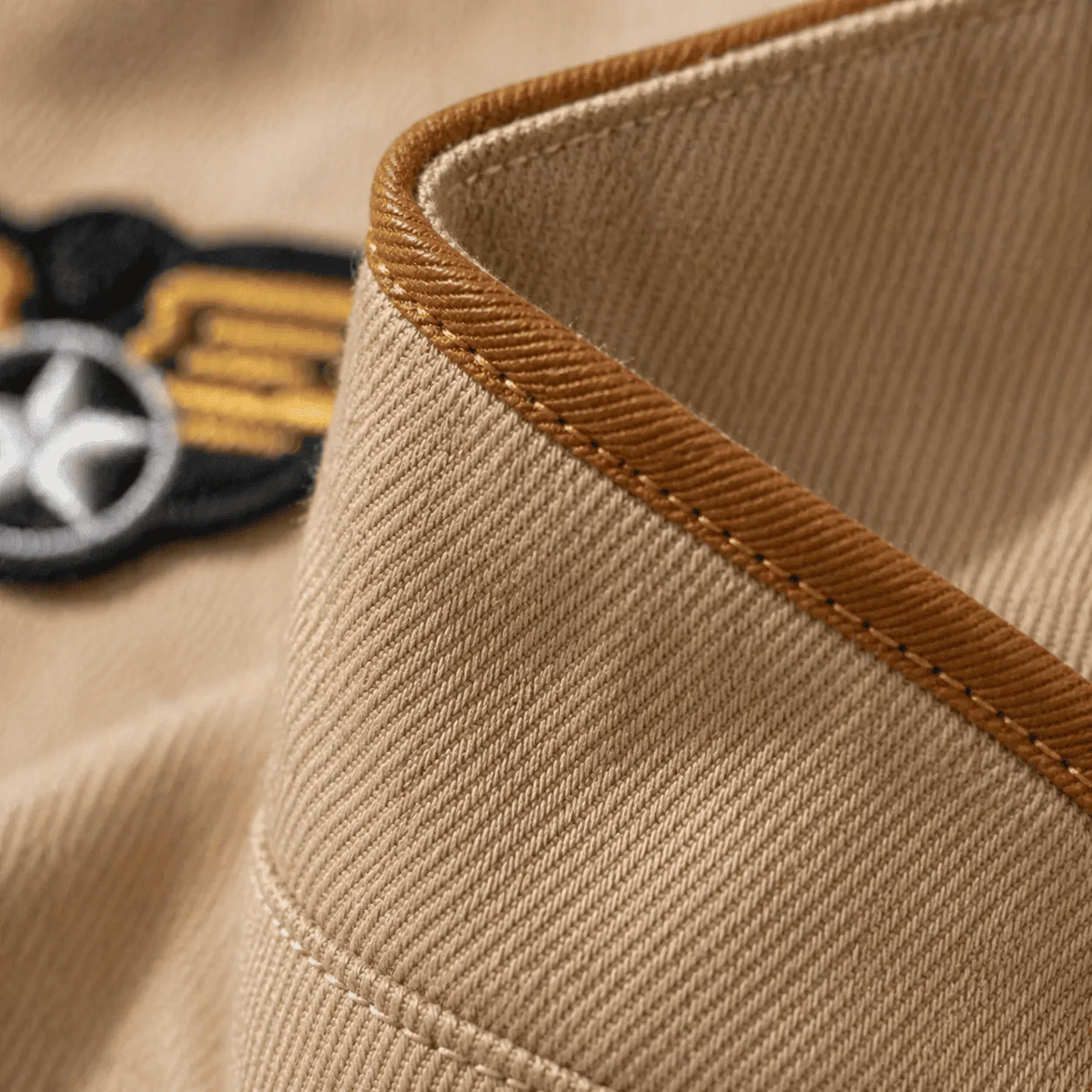Detalhe macro do viés biaset em caramelo acabando a borda da gola do Macacão Aviador em sarja bege, com pespontos caramelo e bordado de asas parcialmente visível