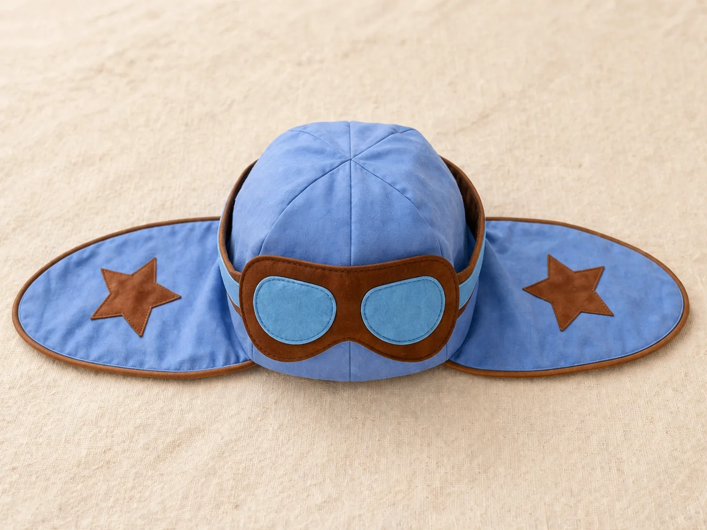 Touca aviador em tricoline azul claro com óculos em feltro de lente azul e contorno marrom · 2 estrelas marrom-cobre nas laterais · tapa-orelha estendido tipo asas
